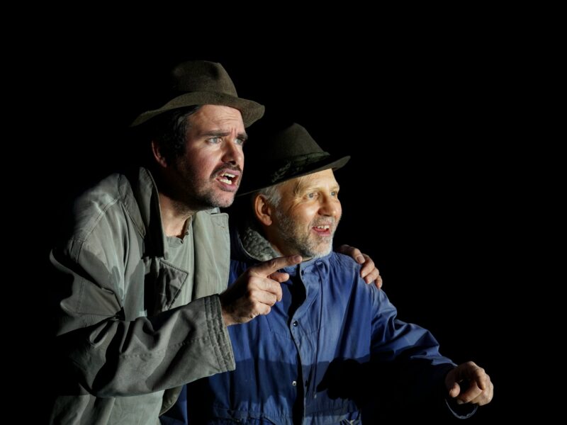Szenenfoto aus „Warten auf Godot“: Klaus Reitberger als Wladimir und Franz Osl als Estragon stehen oder sitzen neben einem kahlen Baum und warten auf die Figur Godot.